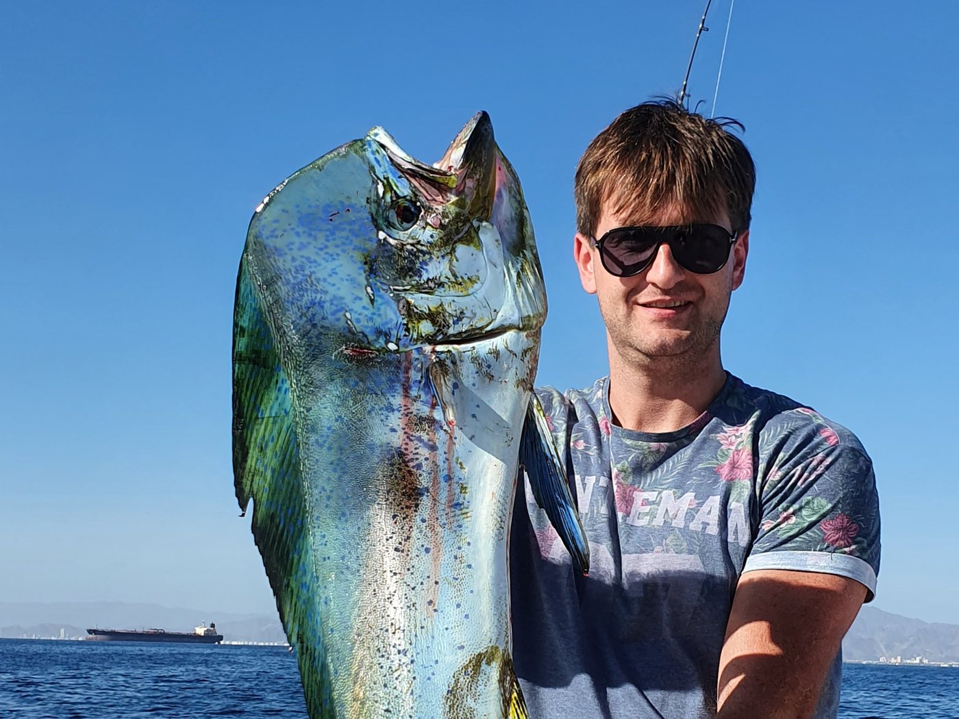 A guest at the rail holding a large dorado, Fujairah coast behind
